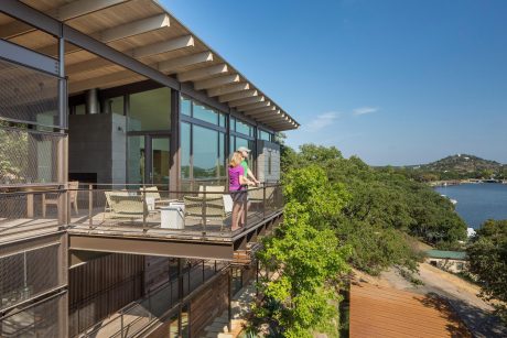 Modern structure with wooden beams, glass walls, and decking overlooking a lake surrounded by lush vegetation.