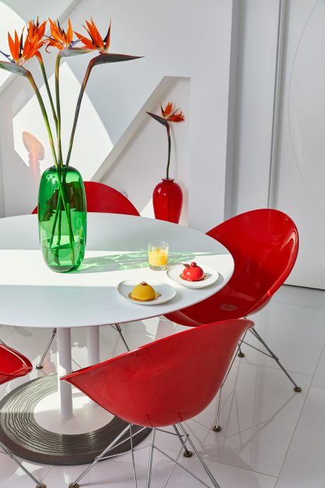 A modern dining area with vibrant red chairs, geometric table, and stylized flower vase.