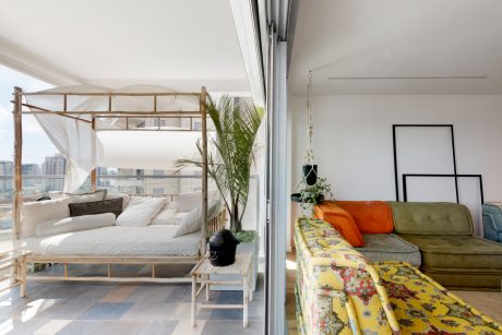 A modern, open-concept interior with a cozy bed, vibrant sofa, and large window offering cityscape views.