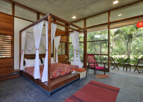Rustic wooden framed bed with canopy suspended in airy, glass-walled room overlooking lush greenery.