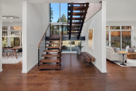 Modern interior with open staircase, hardwood floors, and large windows overlooking trees.
