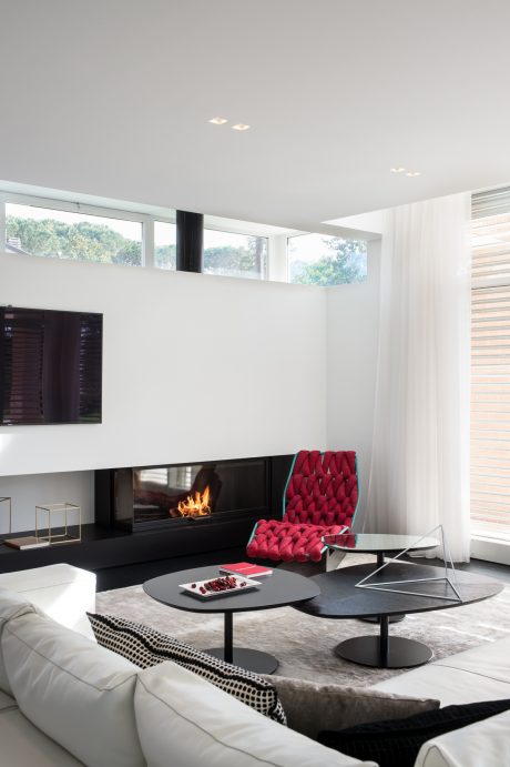 A modern, minimalist living room with a sleek fireplace, tufted red chair, and black glass coffee tables.