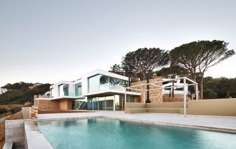 Modern villa with sleek glass walls, stone exterior, and an infinity pool surrounded by lush greenery.