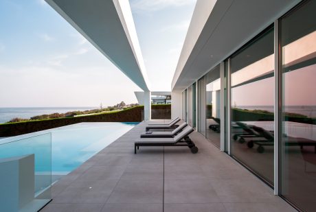 Sleek, modern architecture with glass walls, infinity pool, and lounge chairs overlooking the ocean.