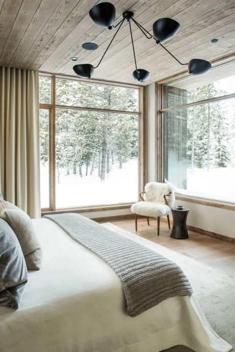 A cozy winter bedroom with a modern chandelier and large windows overlooking a snowy forest.