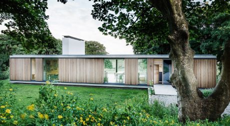 A modern, single-story wooden structure surrounded by lush greenery and a flowering meadow.