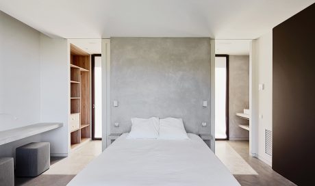 Minimalist bedroom with concrete accent wall, built-in shelving, and large window openings.
