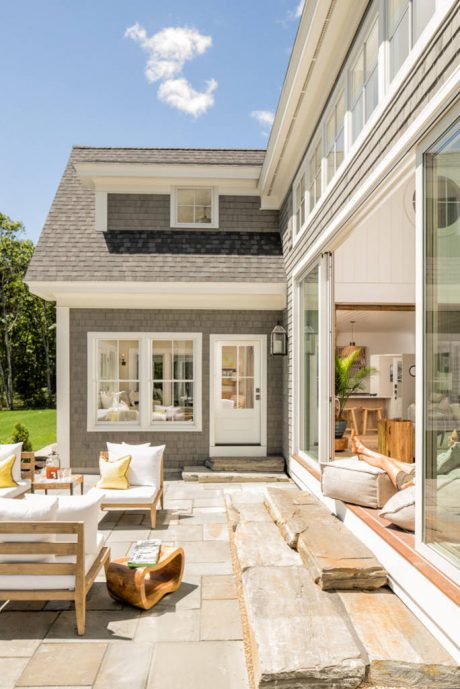 Exterior view of a modern country-style home with gray shingles, wood accents, and large windows opening to a patio.