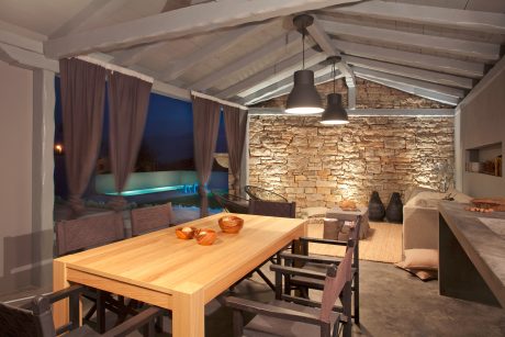 Rustic stone wall, wooden furniture, and illuminated pool create a cozy ambiance.
