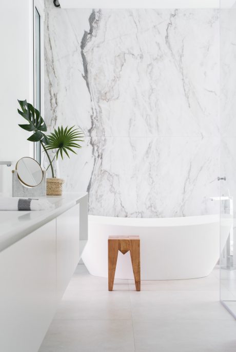 A modern bathroom with a white bathtub, marble walls, and a wooden stool.