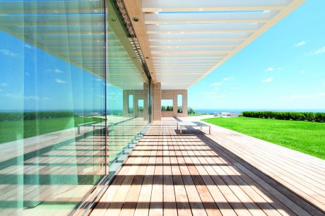 A modern, glass-walled structure with a wooden deck, set against a blue sky and lush greenery.