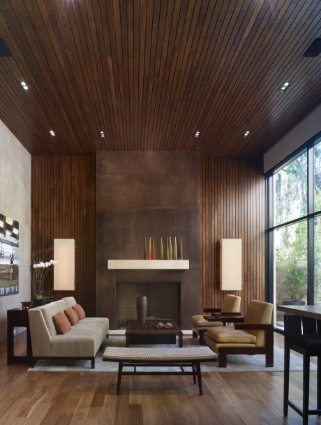 Warm, modern living room with wood-paneled ceiling, concrete fireplace, and large windows.