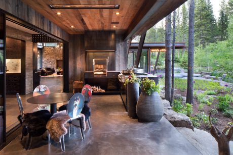 A cozy rustic interior with wooden beams, stone fireplace, and outdoor patio overlooking a lush forest.
