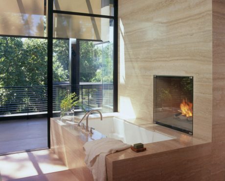 Luxurious bathroom with large soaking tub, fireplace, and floor-to-ceiling windows overlooking nature.
