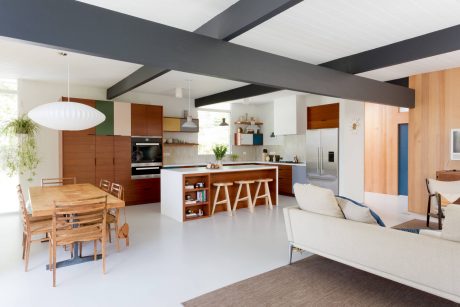 Open-concept kitchen and living room with modern wooden cabinetry, gray beams, and white floors.