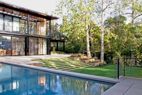 A modern glass house surrounded by lush greenery and a tranquil swimming pool.