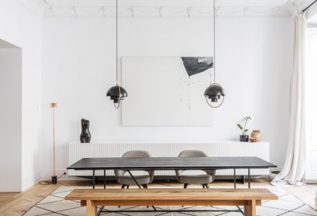 A modern, minimalist dining room with a long wooden table, pendant lamps, and abstract artwork on the wall.