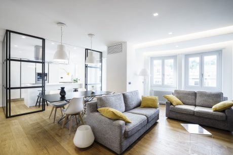Spacious, modern apartment with open-concept living, dining, and kitchen areas featuring minimalist black-framed glass walls and natural wood floors.