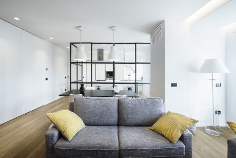 Minimalist living room with open-concept kitchen, glass partition, and modern lighting.