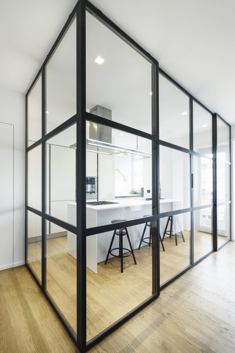 A modern kitchen behind large black-framed glass panels, with sleek white cabinetry and stools.