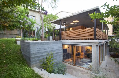 A modern residential building with a partially underground floor, covered deck, and lush greenery.