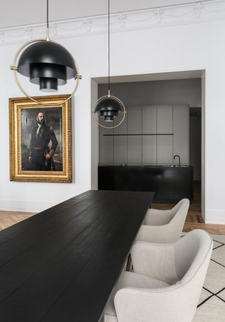 The image showcases a modern, minimalist kitchen design with sleek black countertops, gold-accented pendant lights, and a statement portrait in a gilded frame.