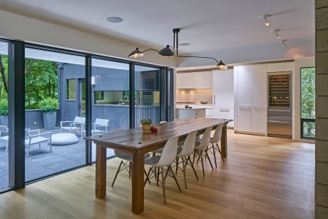 A modern open-concept kitchen and dining area with large windows, wooden furniture, and sleek appliances.