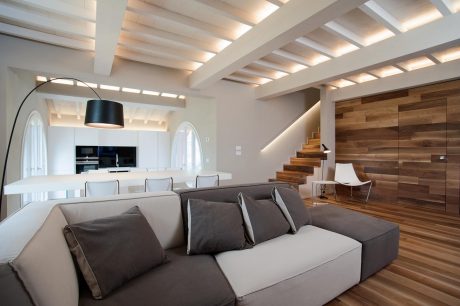 A modern, open-concept living space with exposed wooden beams, a gray sectional sofa, and a wooden staircase.