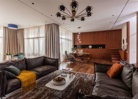 Spacious modern living room with wooden walls, large windows, and a striking brass chandelier.