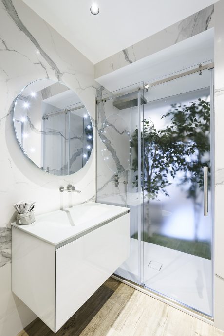 A sleek, modern bathroom with marble walls, LED-lit vanity mirror, and a glass shower with a stunning outdoor view.