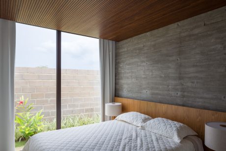 Minimalist bedroom with concrete walls, wooden ceiling, and large window overlooking greenery.