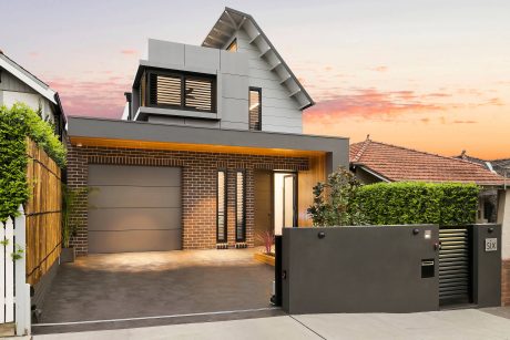 Modern two-story house with gray siding, brick accents, and angled roofline. Garage door and entry gate create a sleek, contemporary exterior.