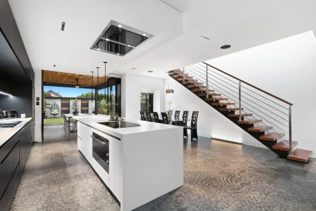 Modern open-concept kitchen with sleek white cabinets, black appliances, and wooden staircase leading to upper level.