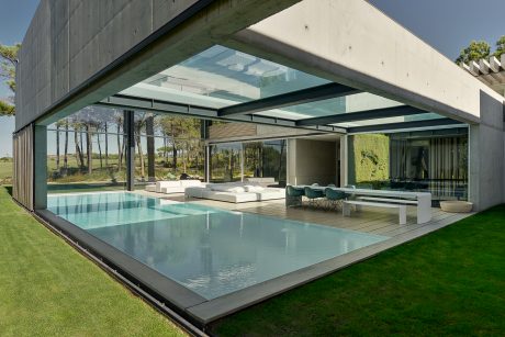 Expansive modern home with glass walls, a swimming pool, and lush green surroundings.