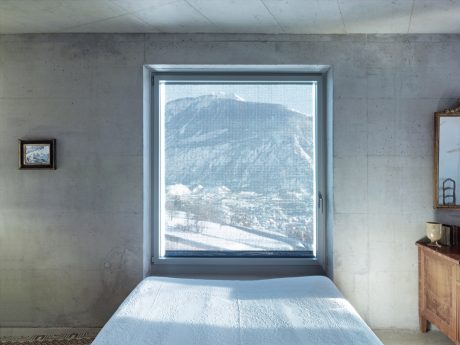 A concrete room with a large window framing a snowy mountain landscape, complemented by minimal furniture.