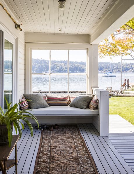 A cozy lakeside porch with a built-in bench, patterned rug, and expansive window offering scenic views.