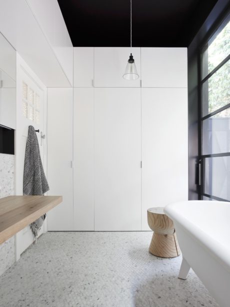 A modern, minimalist bathroom with sleek white cabinets, a wooden bench, and a pendant light fixture.
