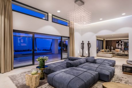 An expansive, modern living room with a large, plush sectional sofa, crystal chandelier, and panoramic windows offering a scenic nighttime view.