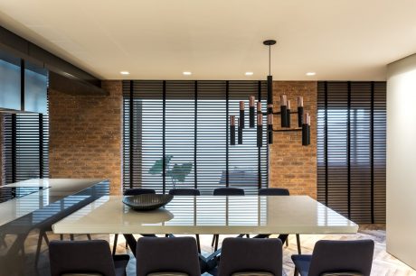 A modern dining room with exposed brick walls, sleek black window blinds, and a large pendant light fixture.