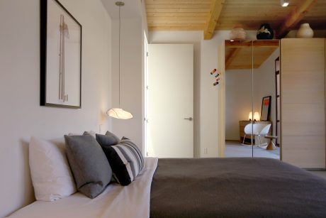 A cozy bedroom with wooden ceiling beams, minimalist decor, and a modern lighting fixture.