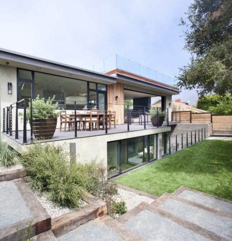 A modern, open-concept home with glass walls, a wooden porch, and a lush garden.