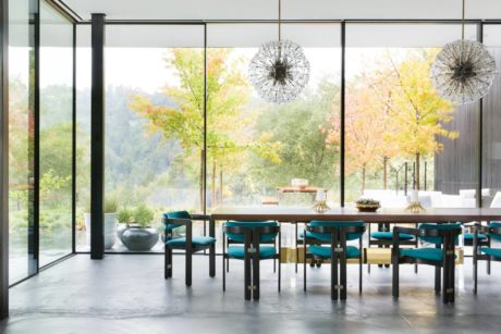 Sleek glass walls frame autumnal landscape; dining table, teal chairs, and modern light fixtures.