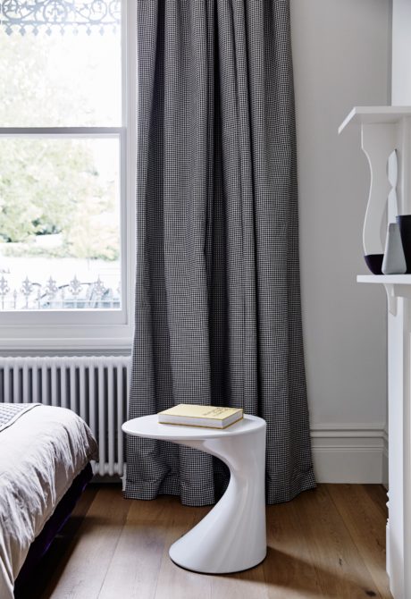 A cozy bedroom with black and white checkered curtains, a white side table, and wooden floors.