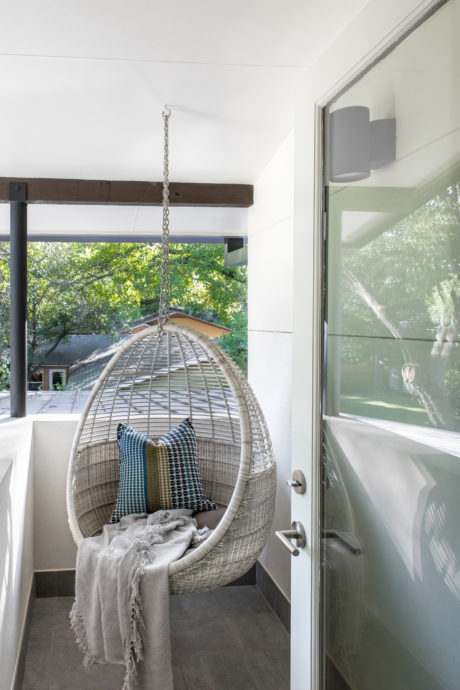 A cozy hanging wicker chair in a modern, light-filled room with large windows overlooking greenery.