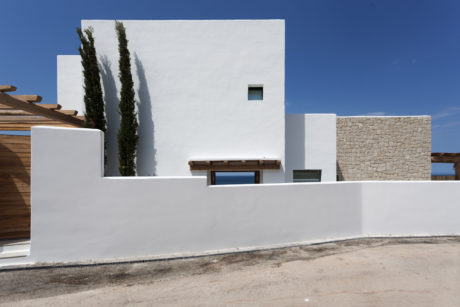 Minimalist white villa with stone walls, wood beams, and tall cypress trees.