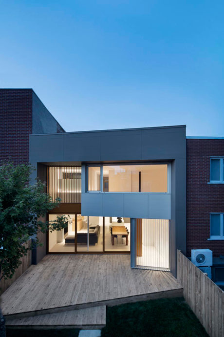 A modern residential structure with a glass-walled interior, wooden deck, and contrasting brick exterior.