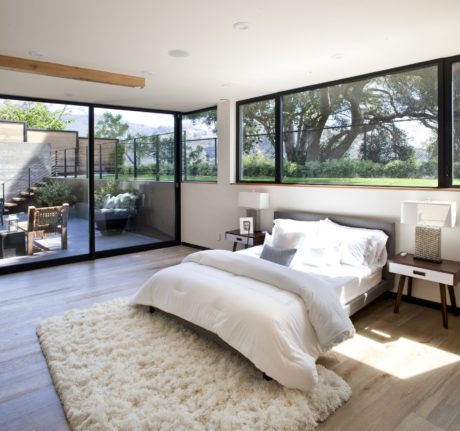 Spacious bedroom with large windows overlooking lush greenery, minimalist decor, and plush rug.