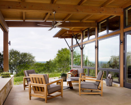 Covered patio with wooden furniture and a view of greenery through large windows.