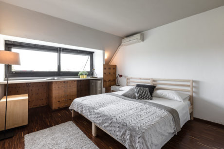 A cozy, minimalist bedroom with wooden furniture, plush bedding, and a large window.