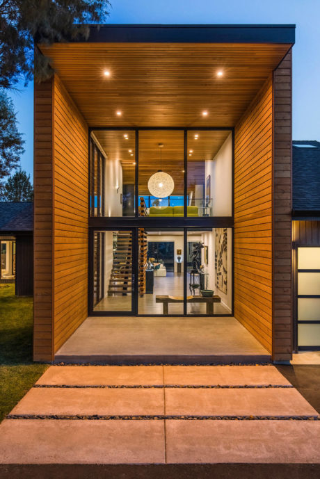 Modern house entry with wood siding, glass doors, and illuminated interior at dusk.